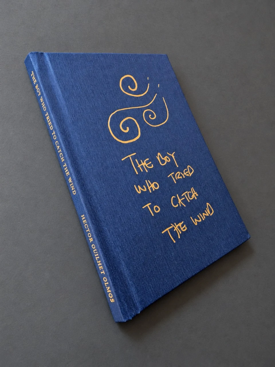 The blue clothbound book on a grey surface, with the handwritten title and wind motif in gold on its cover and spine
